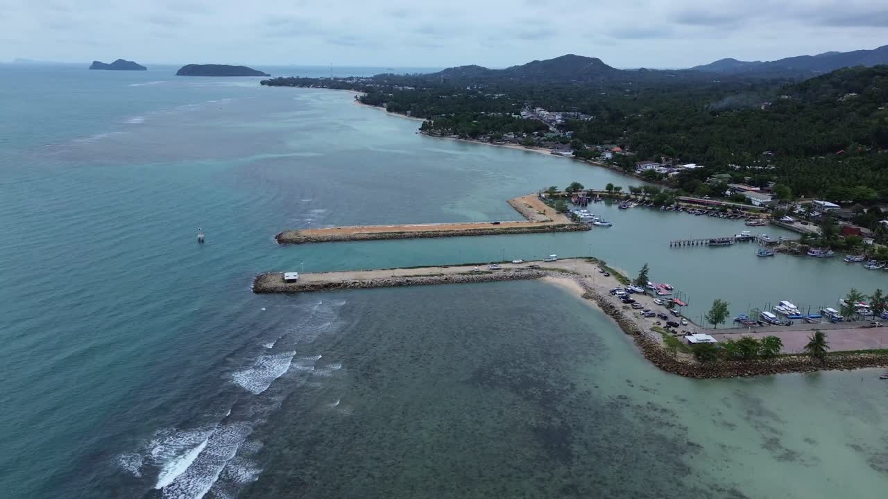 Aerial drone footage showing Baan Tai Pier and the surrounding coastal town on Koh Phangan island, Surat Thani, Thailand, with tropical hills, beach shoreline, and blue sky