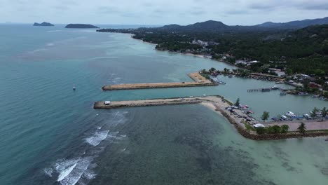 aerial drone footage showing baan tai pier and the surrounding coastal town on koh phangan island, surat thani, thailand, with tropical hills, beach shoreline, and blue sky