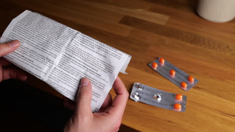 manos sosteniendo la hoja de guía de medicamentos con un blister de tabletas de medicamentos naranjas sobre una mesa de madera