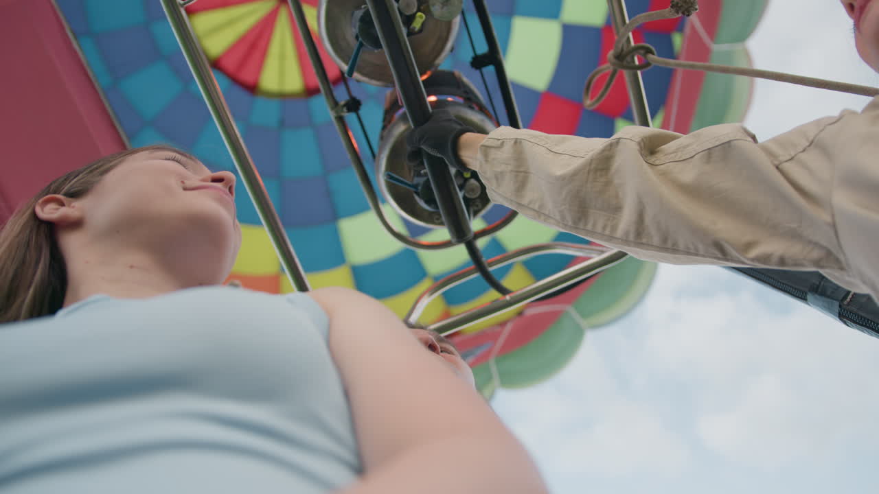 Gloved woman gripping hot air balloon burner lever beneath vibrant envelope panels while friend beside her looks up with a smile, evening light casting warm glow on faces and colorful fabric above