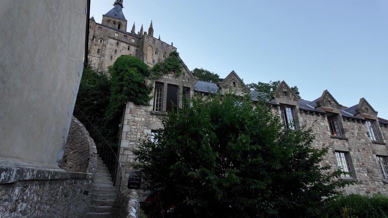 Passeggiata suggestiva lungo le strette strade in pietra sotto l'Abbazia di Mont Saint-Michel, che rivela imponenti mura difensive, blocchi di granito erosi e la drammatica architettura medievale.