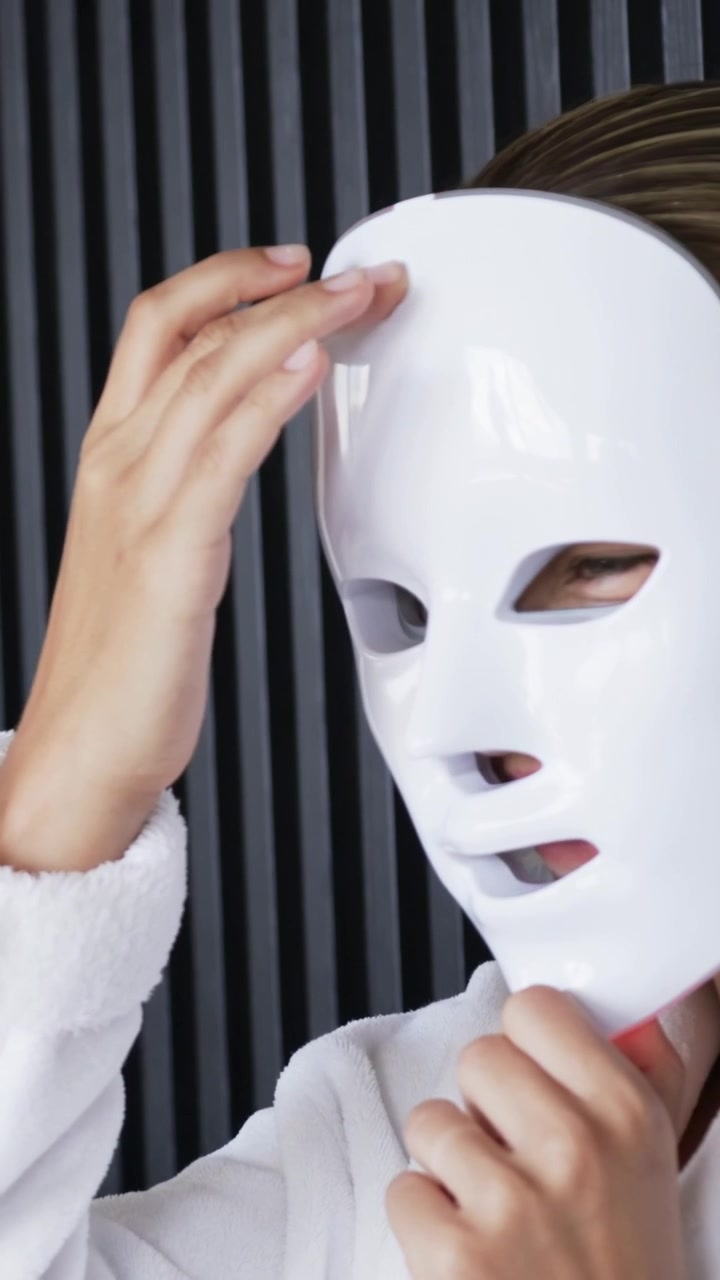 Vertikales Video: Frau greift nach LED-Maske, passt Maske für Spa an, während rote LEDs an schwarzer Wand leuchten.