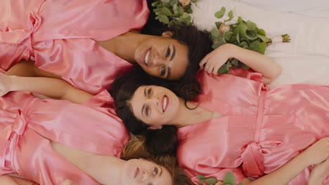 top view of group of multiethnic female friends and bride dressed in pink and white silk nightdresses holding hands with bouquets and laughing while laying on floor 3