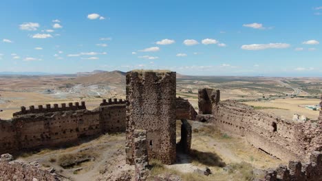 flight-in-a-ruined-castle-with-its-walls-and-towers-seeing-the-main-tower-with-a-panning-of-the-camera-it-is-in-the-middle-of-the-fortress-and-with-crop-fields-and-a-town-on-a-summer-Toledo-Spain