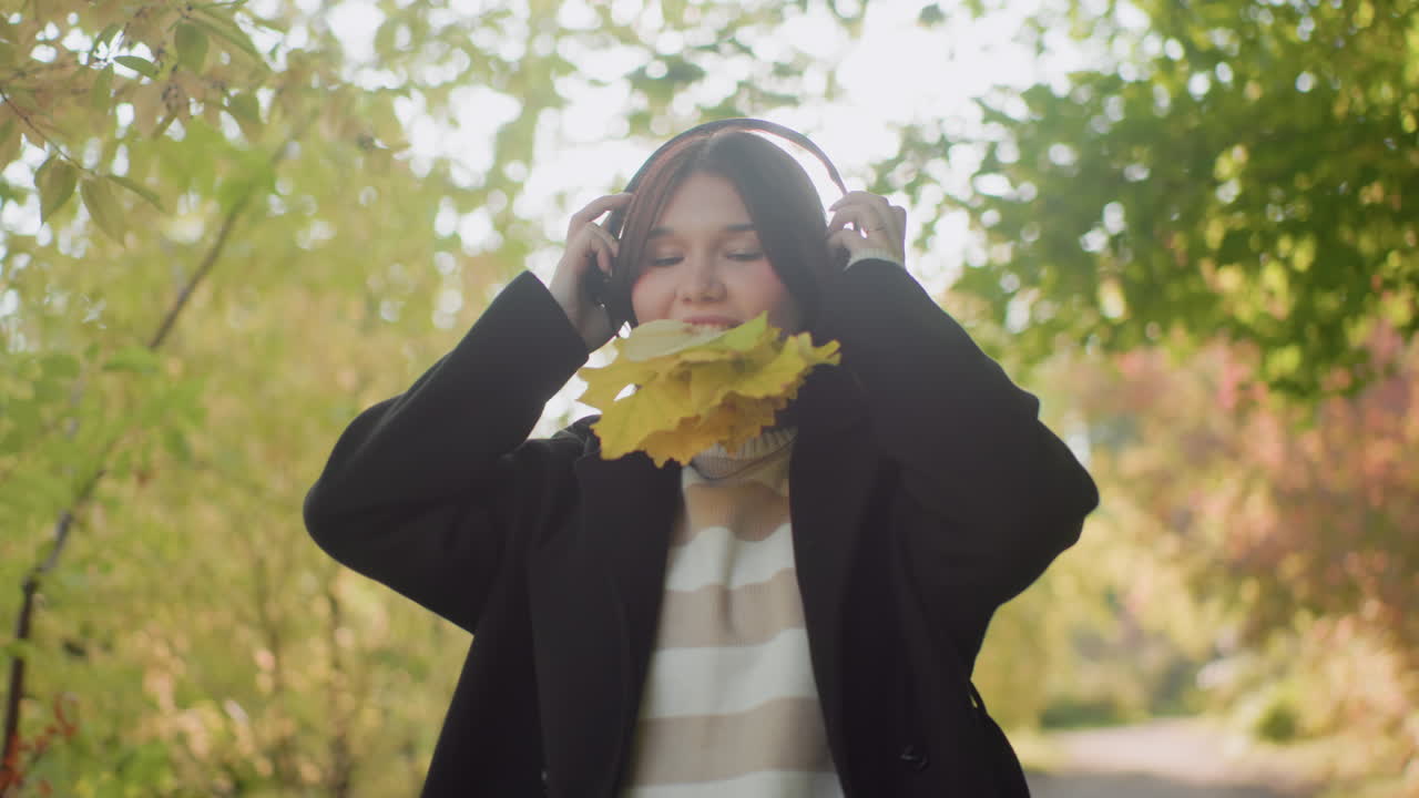 Medium view of autumn lover outdoors holding leaves in mouth while lifting headset to ears, pausing on forest path before continuing relaxed walk, soft sunlight, golden bokeh, cozy sweater