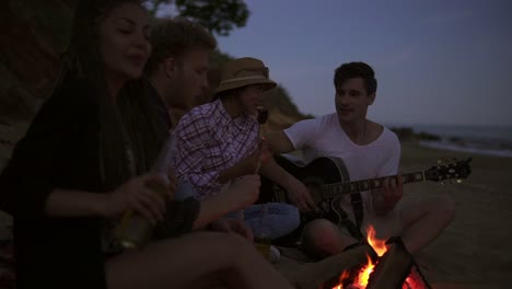 picnic of young people with bonfire on the beach in the evening. cheerful friends singing songs and playing guitar. slow motion