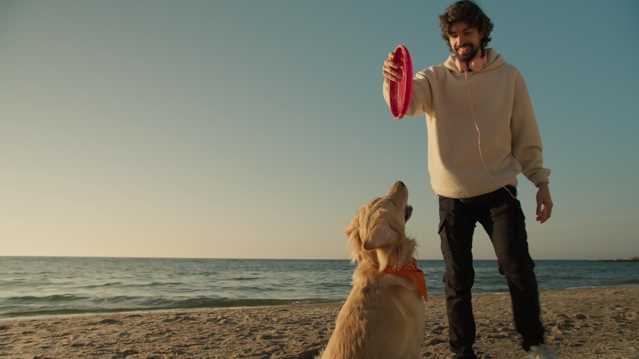Shooting close up of a young guy playing with his dog light coloring with a red toy on the sunny beach in the morning