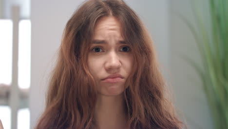 Unhappy-young-woman-touching-and-looking-on-dry-hair-ends-in-mirror-at-bath-room