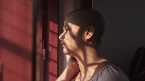 bald indian girl rubs color powder on cheeks on holi looking outside at home due to covid-19 lockdown