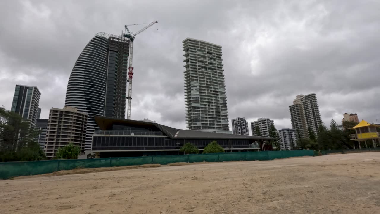 una escena de playa tormentosa con fuertes vientos y olas turbulentas del océano bajo cielos nublados, mostrando edificios urbanos en el fondo