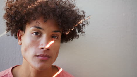 young man with curly hair looking at camera, standing against wall