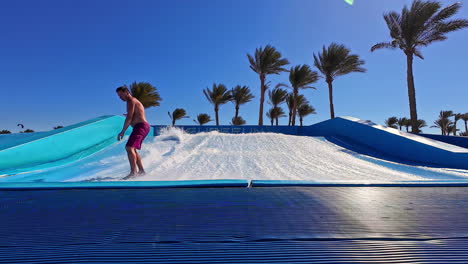 man surfing on artificial wave pool with palm trees in background, sunny day, resort vibes