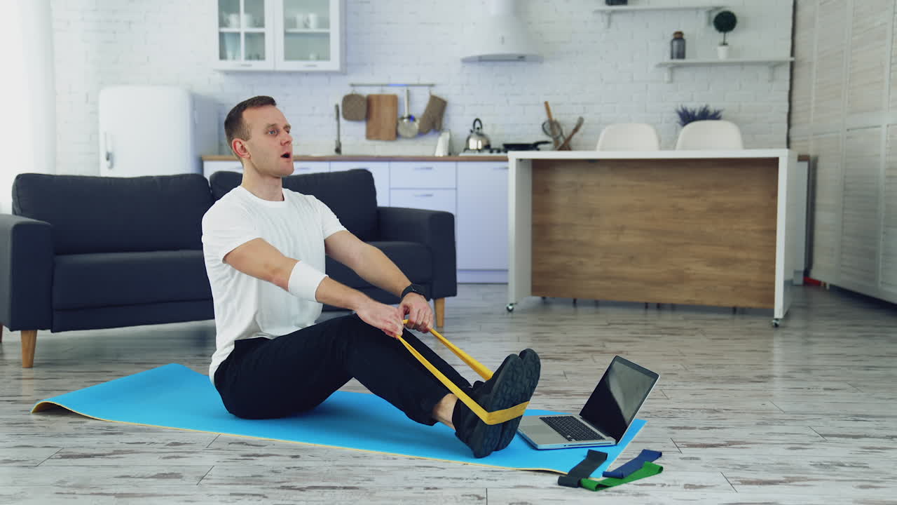Workout at home during quarantine. Young man is doing breath exercises with fitness rubber band on the floor in front of laptop. Sporty lifestyle indoors.