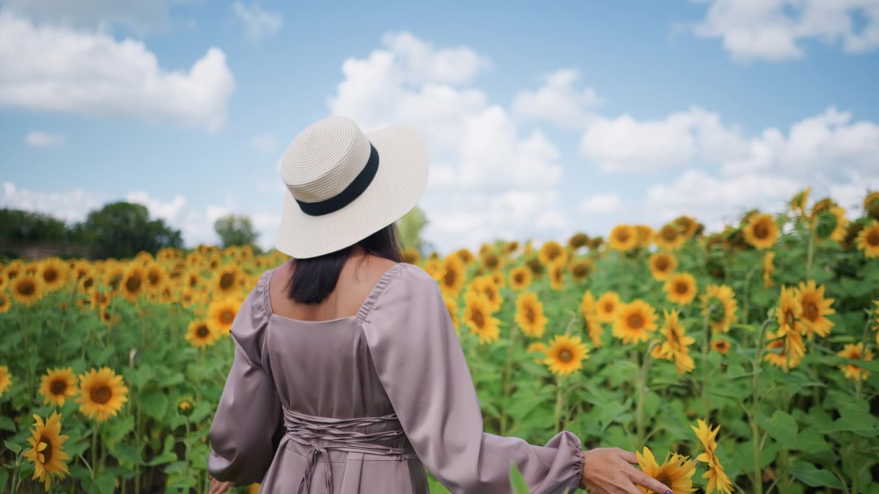 Donna con cappello a tesa larga e abito lungo cammina tra i girasoli toccando delicatamente i fiori con la mano. La scena presenta vivaci fioriture gialle sotto la luce del giorno intensa contro un vasto cielo blu
