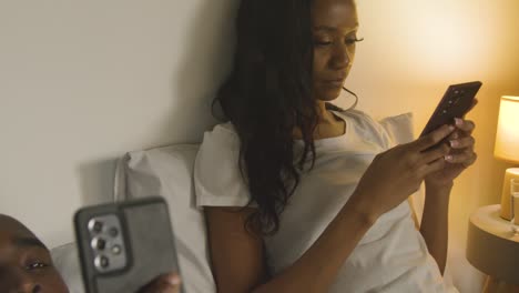 couple with relationship problems at home at night lying in bed both looking at mobile phones 2