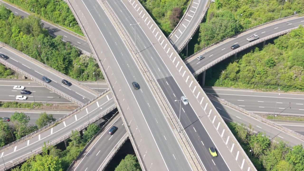 Aerial view of a major highway interchange on a bright day. Multiple cars and vans move along the interconnected roads and ramps, surrounded by green foliage, creating a dynamic urban landscape.