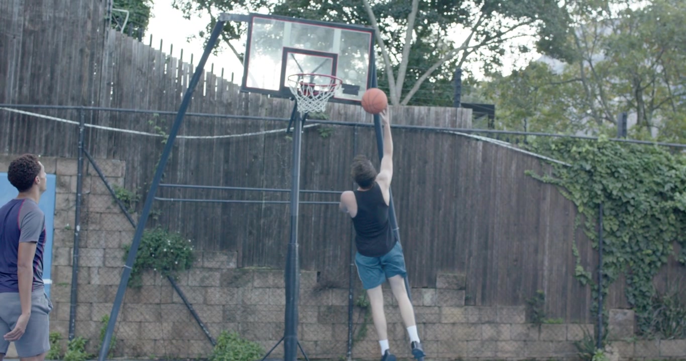 Amigos, un hombre con camiseta sin mangas negra driblando el balón en una canasta de patio trasero, ejecutando un crossover, avanzando para anotar.
