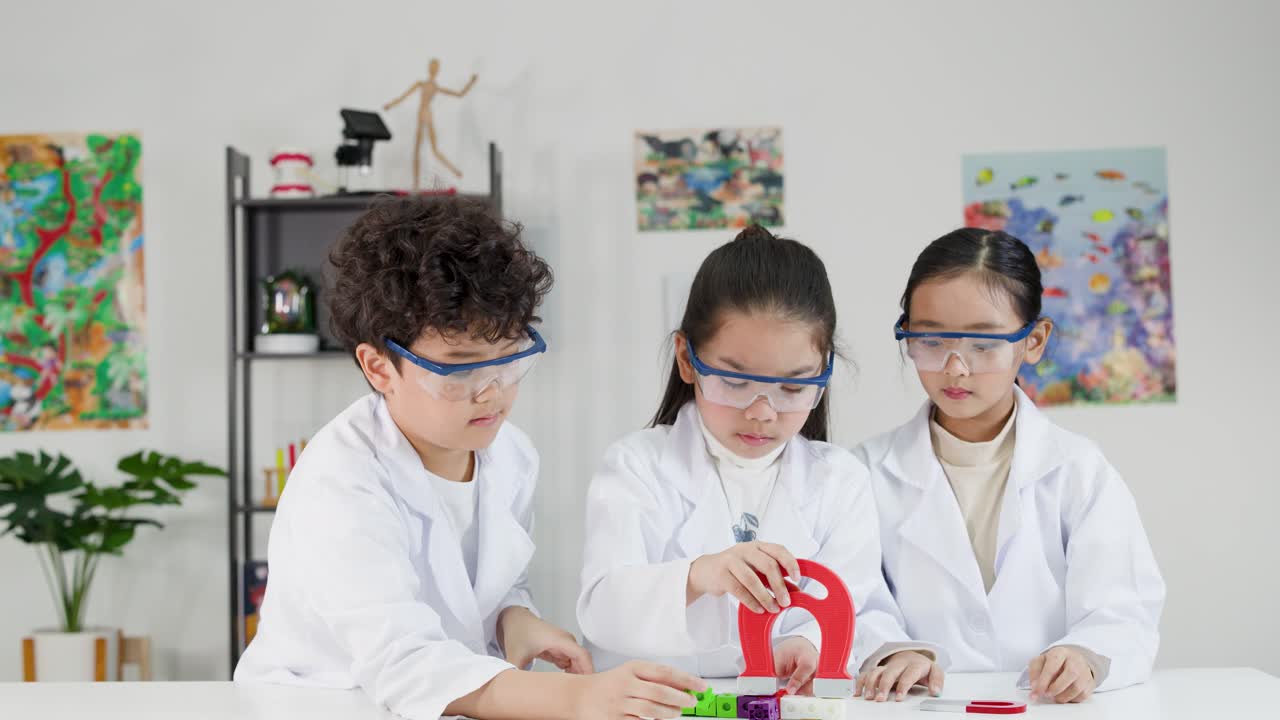 Tiga siswa muda mengenakan kacamata pelindung dan jas laboratorium saat menjelajahi magnetisme di ruang kelas.