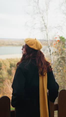 vista trasera de una mujer con el cabello ondulado rojo en un abrigo negro, caminando hacia una valla de madera, descansando su mano en ella mientras disfruta de la vista panorámica de un río y follaje seco