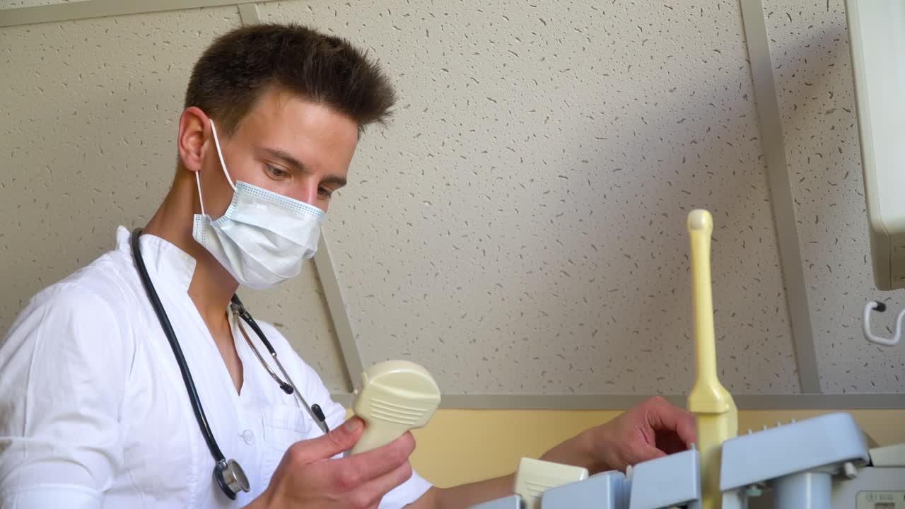 Young doctor in protective mask working on ultrasound device. Doctor adjusts the ultrasound device