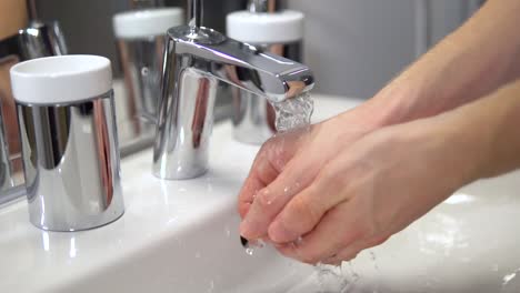 Hombre-Blanco-Previniendo-Enfermedades-Limpiándose-Las-Manos-Con-Agua-De-Grifo-En-Un-Baño-Brillante-Y-Estéril