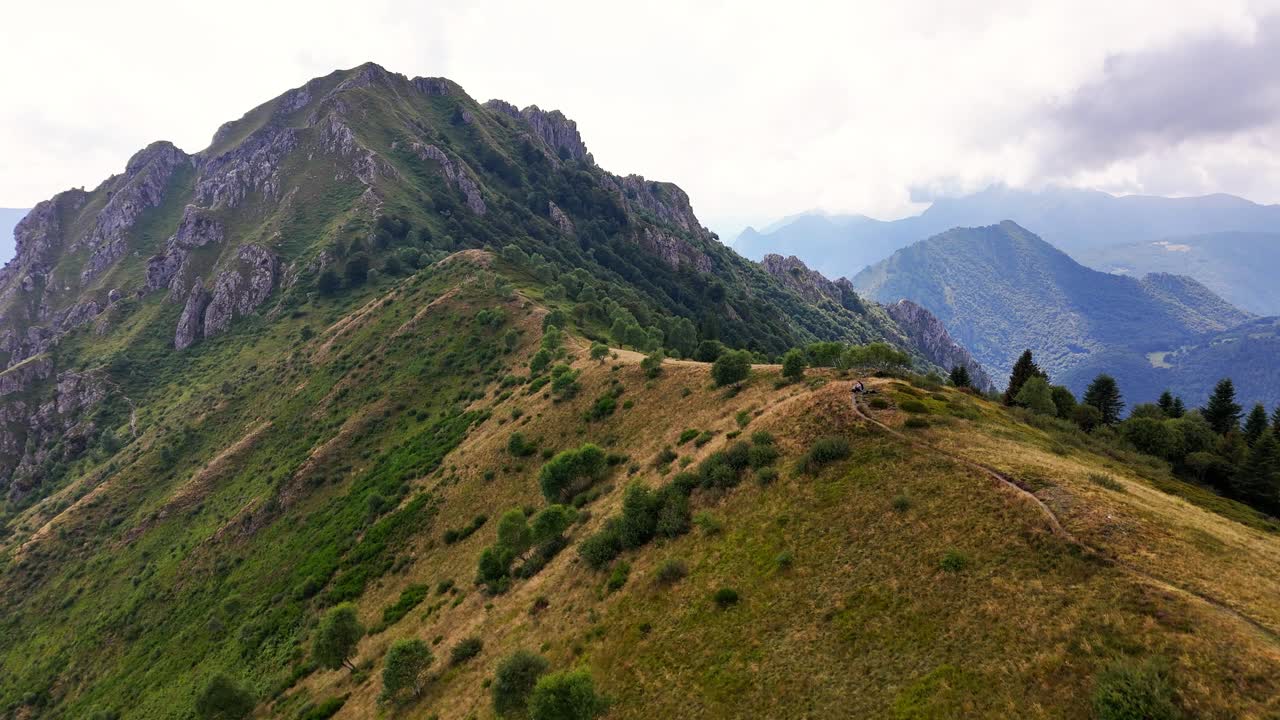Wide aerial view of Cima Monte Grona at Lago di Como in Italy with rugged slopes, winding trail, scattered shrubs and distant layered mountain ranges under soft clouds creating a elevated landscape