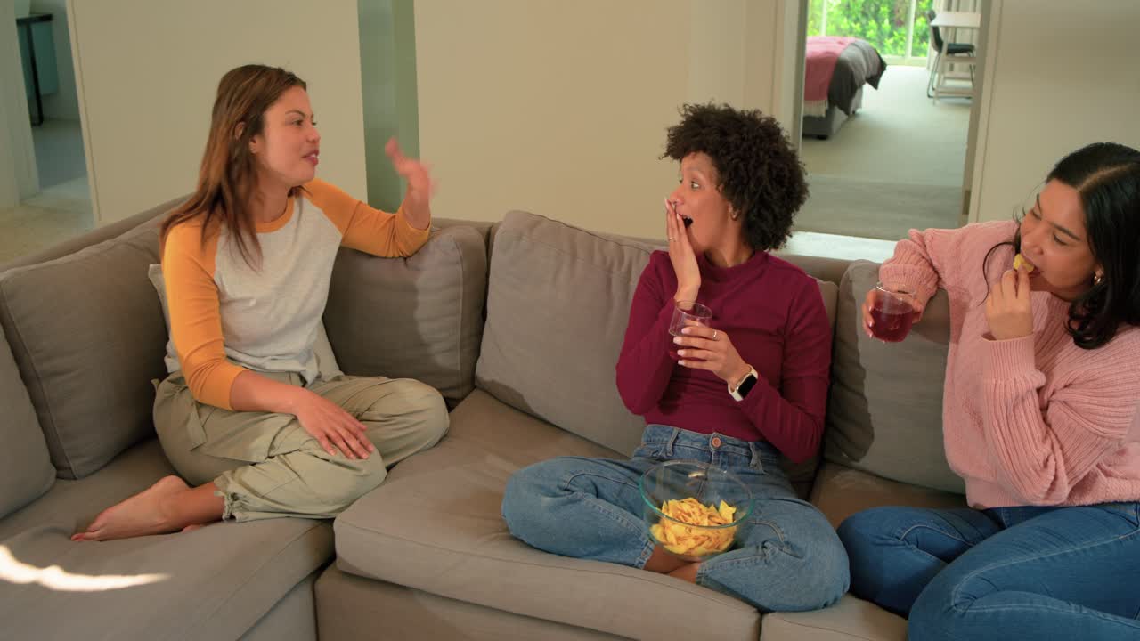 Amigas diversas sentadas no sofá, uma publicando uma história enquanto as outras comem batatas fritas e bebem vinho.