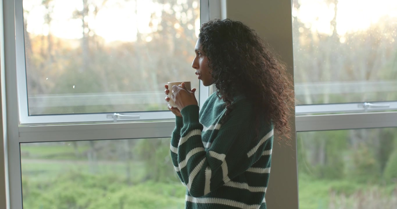 Luce solare che cambia, donna afroamericana alla finestra che alza e abbassa una tazza di ceramica, guardando fuori, con spazio per il testo.