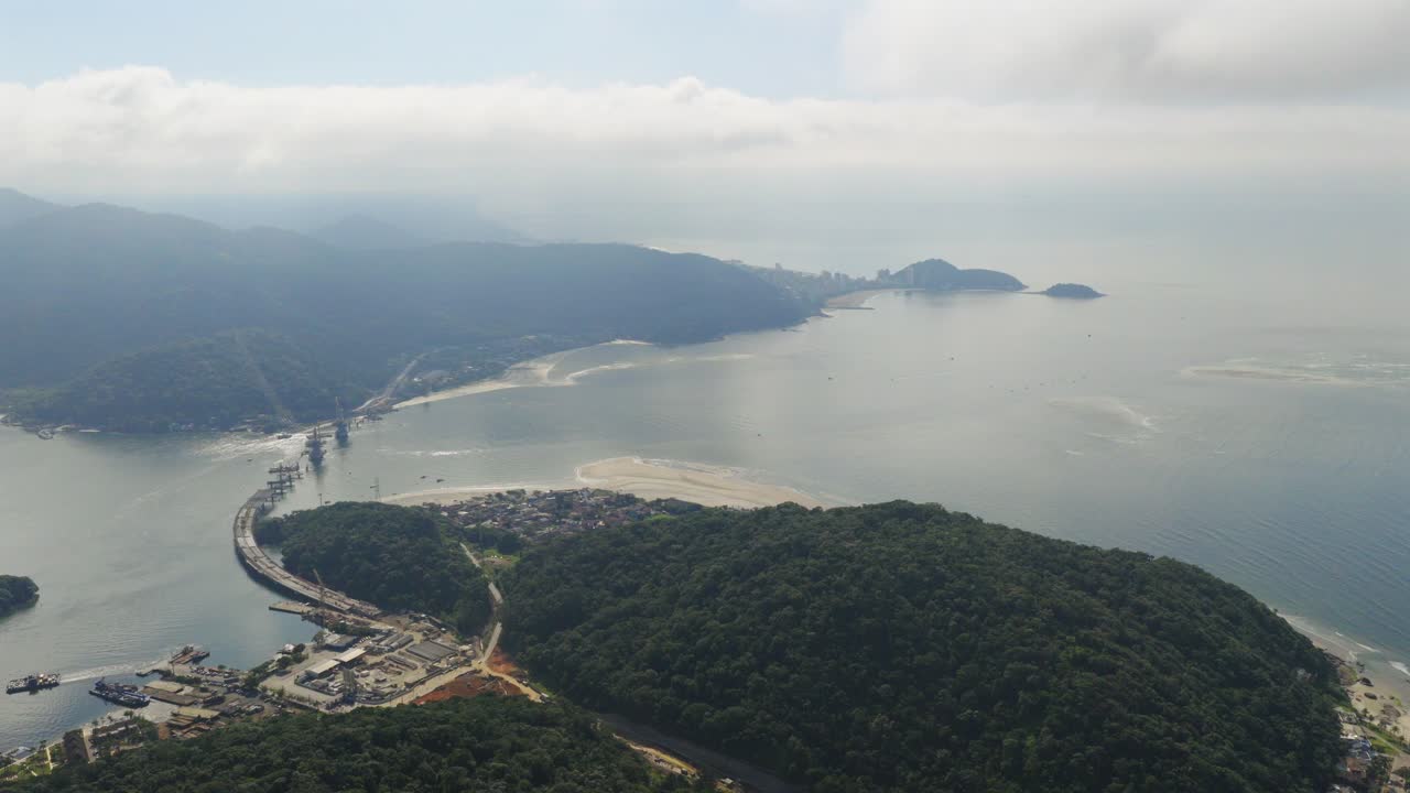 Drone slowly orbits over Guaratuba–Caiobá Bridge construction site, revealing lush hills, coastal towns, and expansive calm bay waters in the State of Paraná, southern Brazil, under cloudy skies