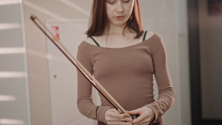 Studente bianco che ispeziona l'archetto del violino prima della pratica, stringendo la vite, controllando il crine, applicando la colofonia, mani attente, custodia aperta sul pavimento, calma concentrazione, preparazione per la prova