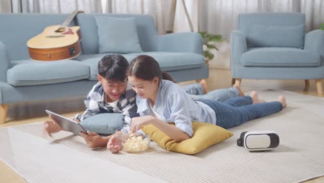 full body of asian teen couple using tablet while lying on carpet on the floor at home. eating snacks watching movie, speaking, and enjoying time together