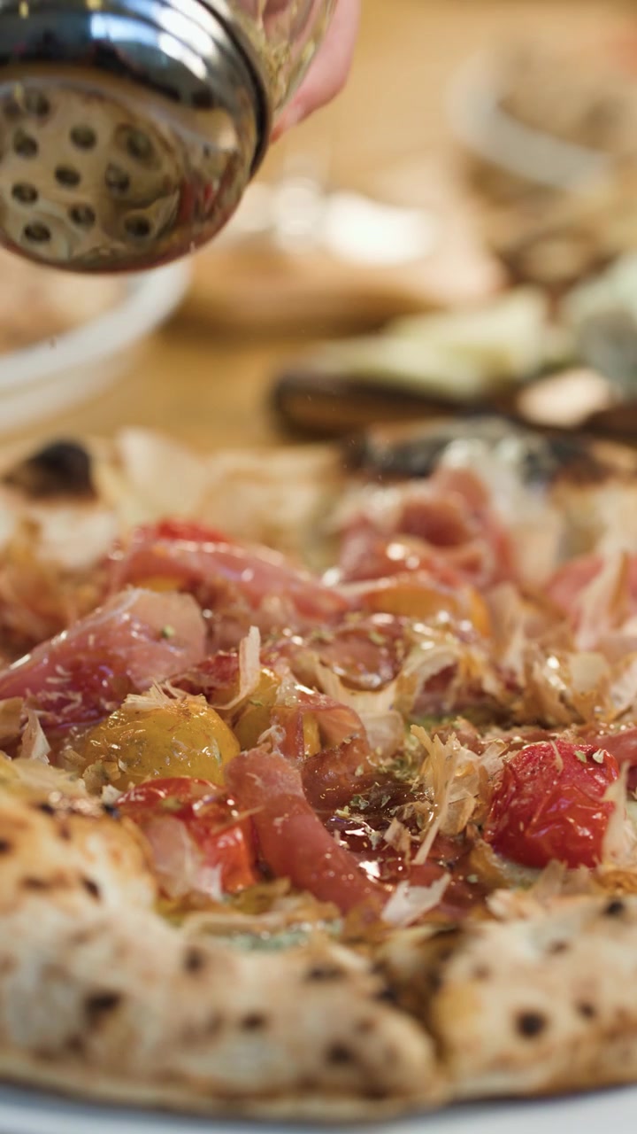 Primo piano di una mano che sparge erbe aromatiche essiccate su una pizza fresca con prosciutto e pomodorini.