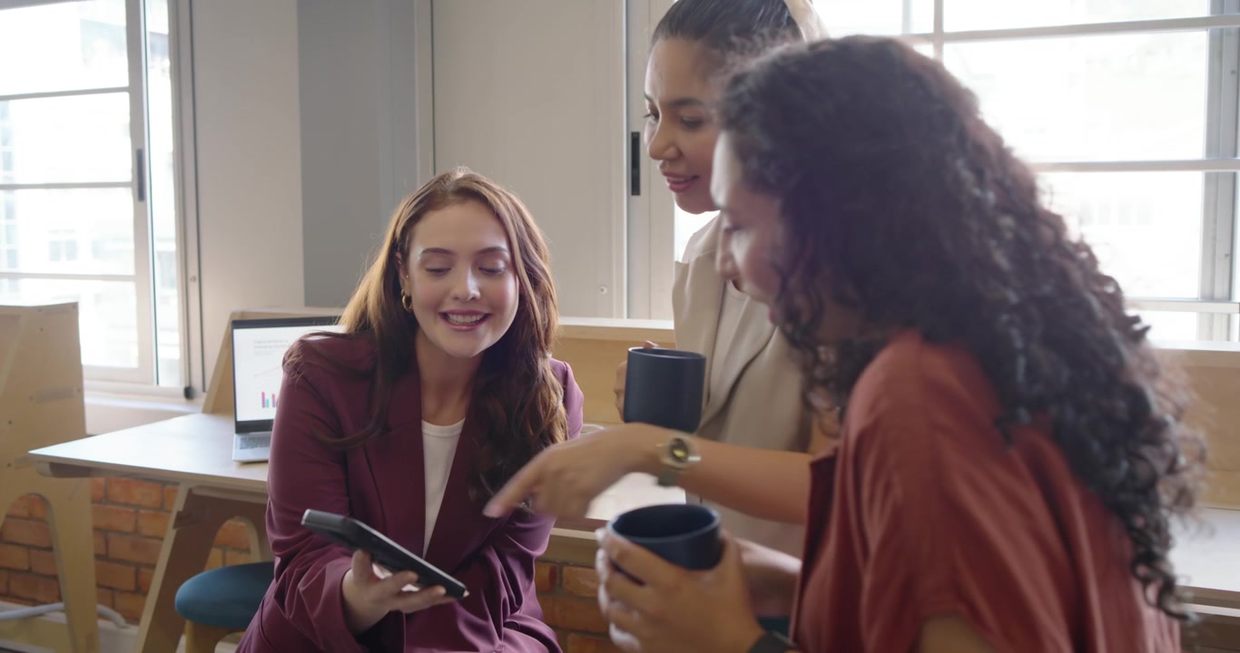 Donne in ufficio, una collega che parla, una donna seduta che scorre il telefono, mostrando agli altri che tengono delle tazze