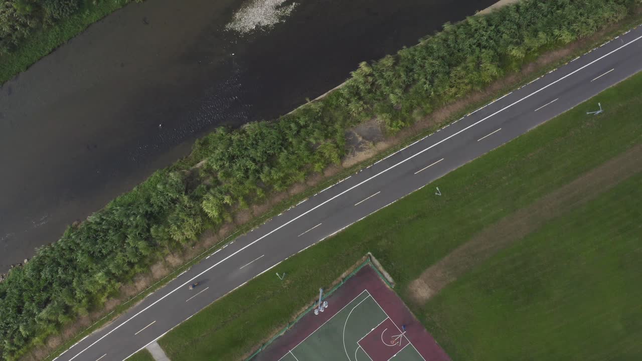 4K drone aerial footage showing a top down view of a straight road beside a river with green grass fields and a basketball court. Iideal for urban planning, travel, sports, or environmental projects