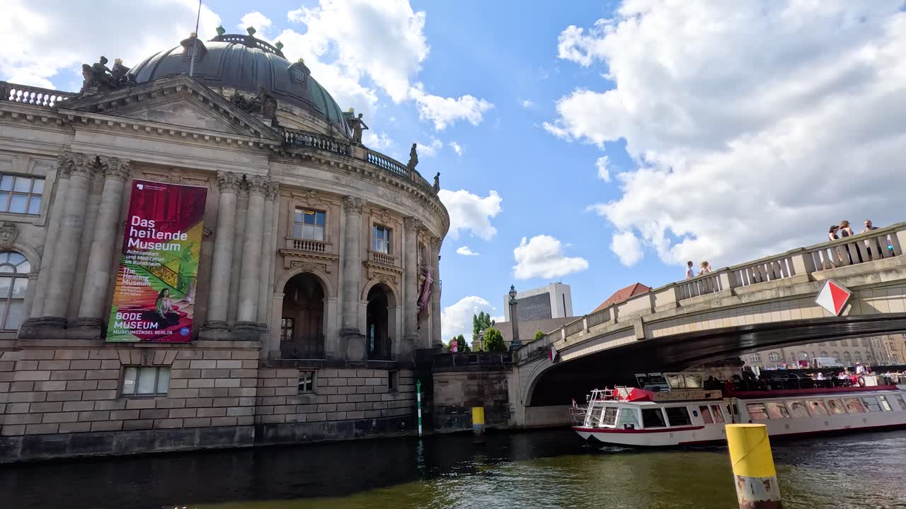 A sightseeing boat glides past a historic riverside museum under a partly cloudy sky, with dynamic camera movement suggesting a river cruise perspective