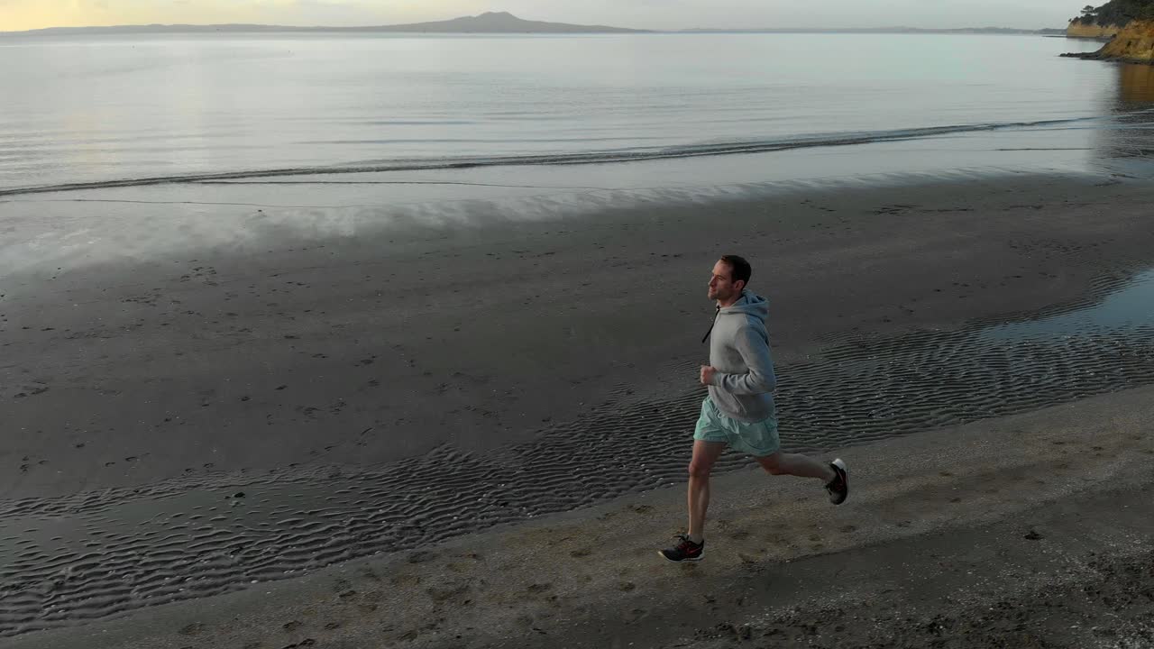 Aerial tracking shot of fit young caucasian man running on a beach in Auckland, New Zealand