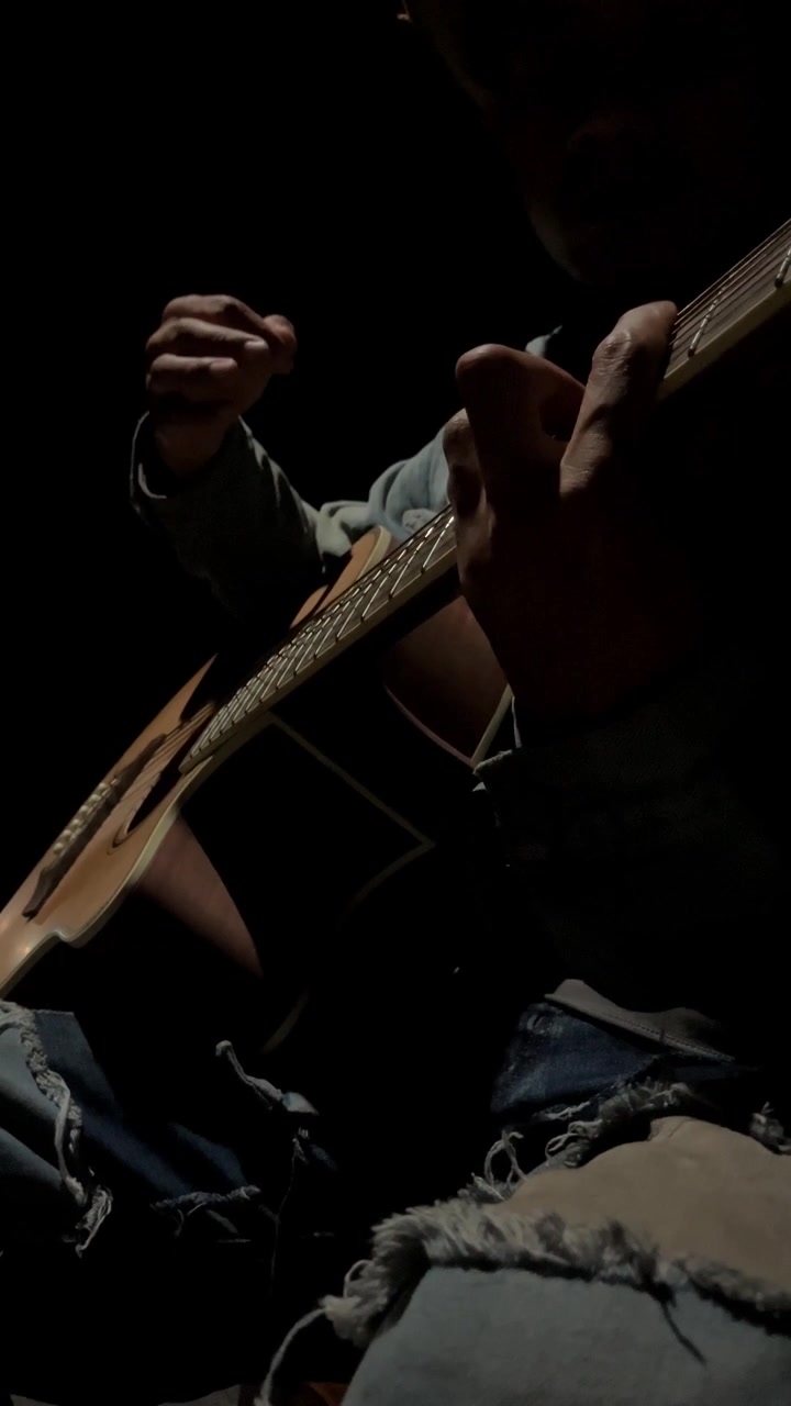Ripresa verticale in 4K al rallentatore da angolazione bassa a livello del suolo di un uomo in jeans strappati che suona una chitarra acustica. L'illuminazione scura, suggestiva e a bassa chiave crea un broll di una performance musicale intima e drammatica