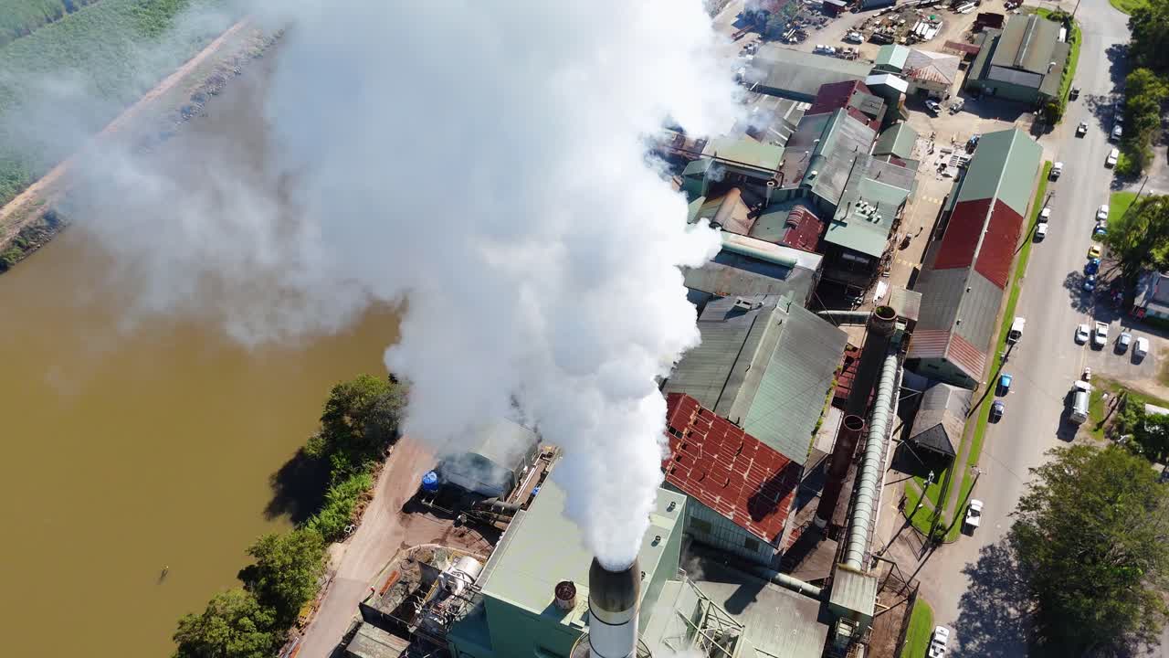 Drone footage captures a factory releasing smoke beside a river, highlighting industrial activity and environmental impact under clear skies