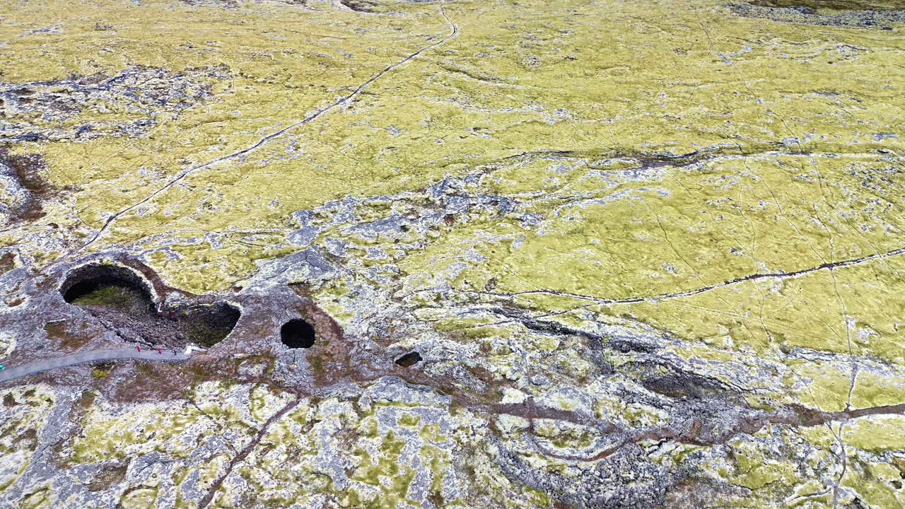 Pemandangan udara drone hamparan luas ladang lava yang tertutup lumut dengan gua vulkanik dan bukaan yang tersebar di seluruh medan. Gunung dan gletser di kejauhan terlihat di cakrawala dataran tinggi Islandia.