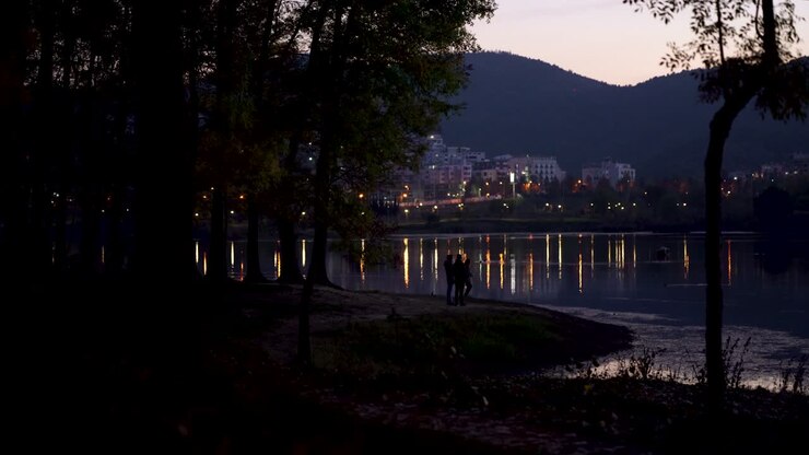 Parco cittadino attorno a un lago calmo che riflette le luci stradali sull'acqua tranquilla al crepuscolo.