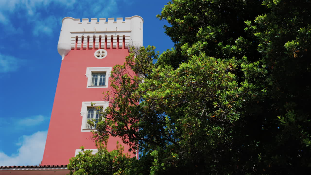 Green tree moving in the wind near the red tower of the Cremat Castle Winery over blue sky
