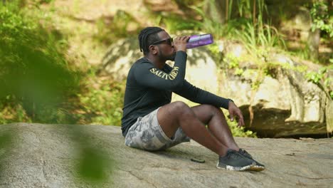 A-man-takes-a-sip-of-water-on-a-giant-rock-in-the-middle-of-the-woods-on-a-bright-sunny-day