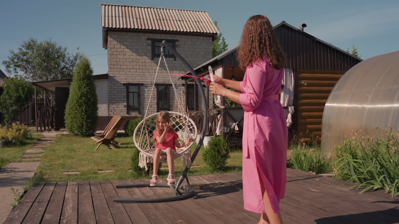 Happy Family Enjoying Porch Swing Together, Mother And Daughter Laugh On Hanging Porch Swing In Warm Sunlight, Cheerful Family Moment With Mother And Child Swinging Outdoors Near Modern House