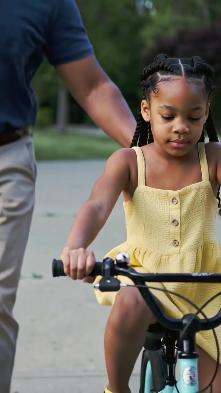 Vertical video: Riding girl in yellow dress learning on road with adult steadying bike navy polo