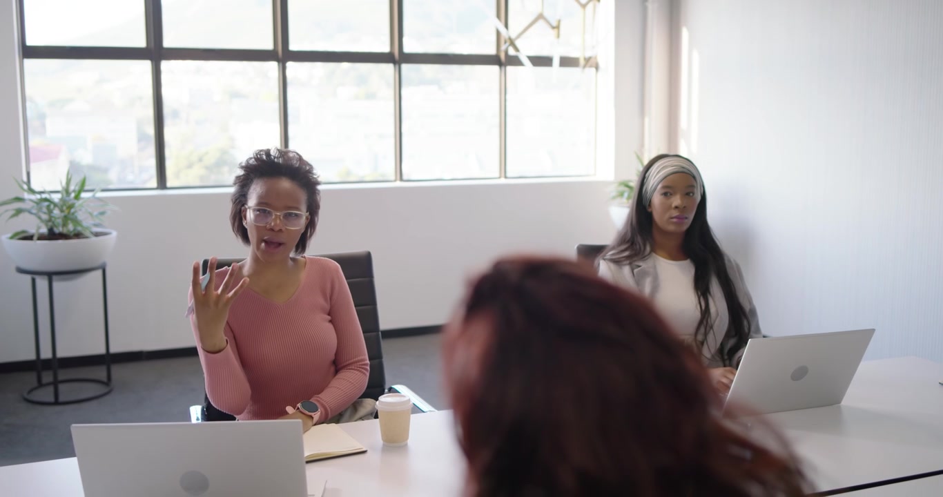 African American women pointing pen speaking at table, laptop user pausing typing