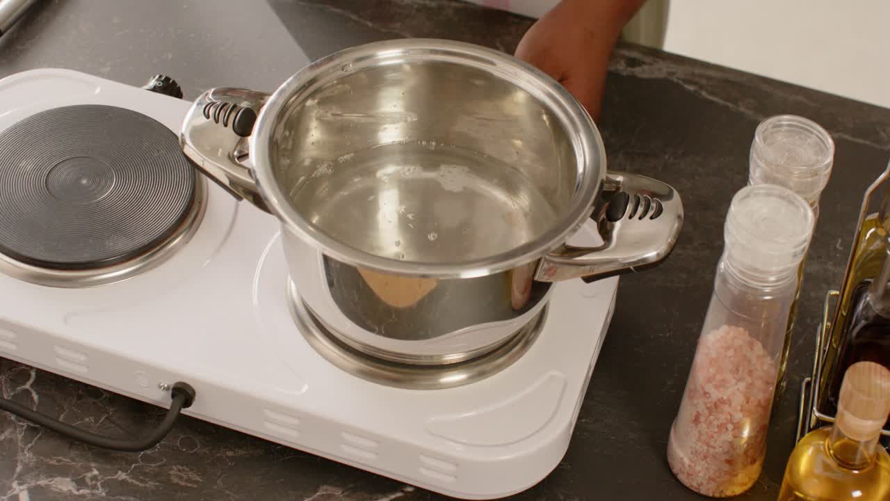 Hombre vertiendo agua de una taza medidora en una olla sobre una placa caliente, alcanzando los controles para calentar el agua.