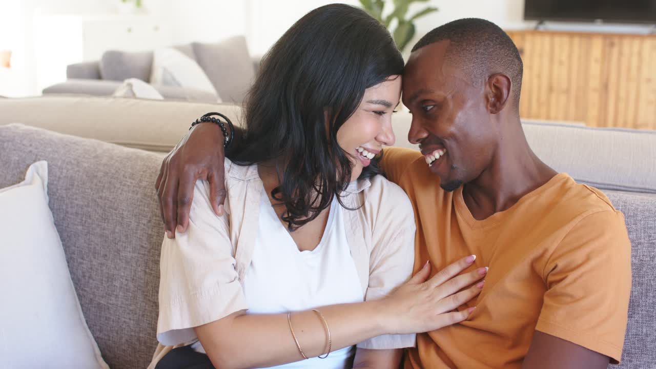 Couple sitting on gray sofa in bright living room, man leaning, draping arm, woman laughing, posing