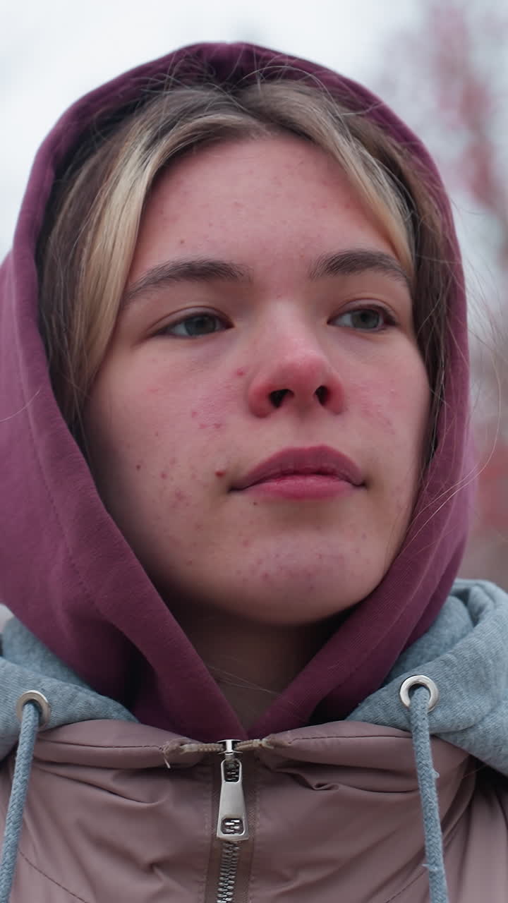 Primo piano di una giovane donna che indossa una giacca invernale con cappuccio, che cammina pensierosa e si guarda intorno con attenzione, in un parco innevato con alberi con bacche rosse e un cielo nuvoloso e tenue.