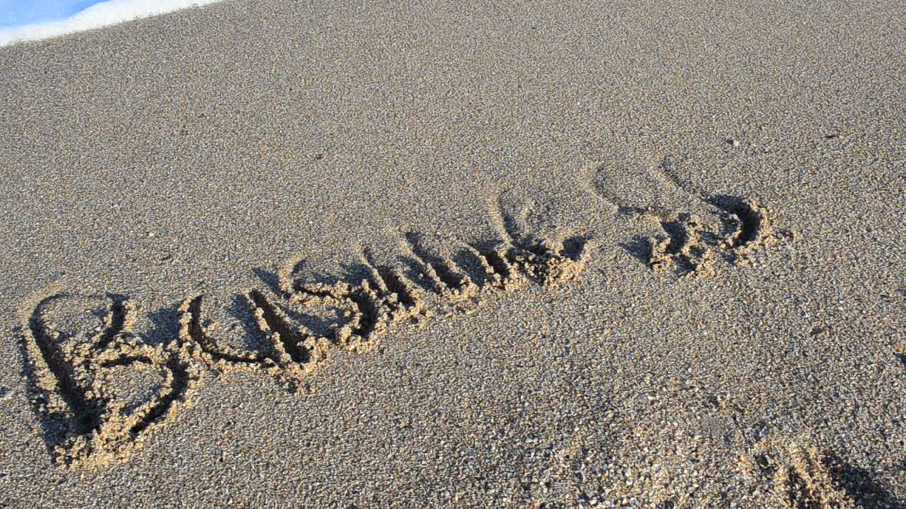 Iscrizione 'business' sulla sabbia. L'iscrizione viene cancellata da un'onda del mare. Riprese sulla spiaggia.