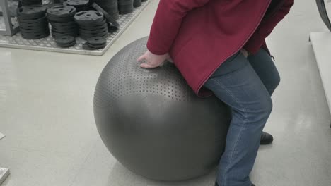 woman in a sports store buys a black fitball. she sits on it and jumps